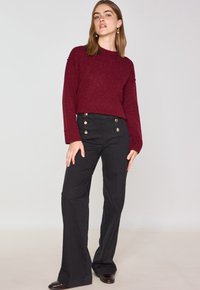 Maglione a maglia color bordeaux con una vestibilità rilassata, abbinato a pantaloni svasati neri con dettagli di bottoni dorati in vita. Texture complessiva liscia.