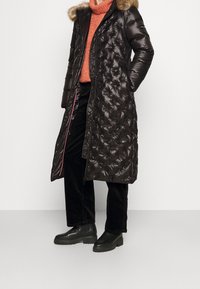 Abrigo acolchado negro con capucha forrada de piel, patrón acolchado, cremallera frontal y forro rosa en contraste, llevado sobre un suéter naranja y pantalones negros.