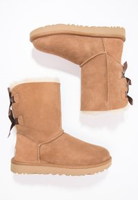 Botas de tobillo de ante castaño con forro de piel suave, suela de goma acanalada y lazos decorativos en los lados. Textura suave, diseño redondeado.