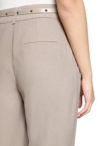 Pantalon beige ajusté avec une texture lisse, une coupe slim, une poche arrière unique et une ceinture contrastée avec des accents métalliques.