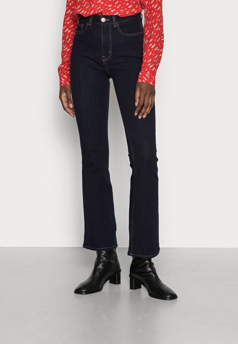 Jean évasé bleu marine taille haute avec des coutures contrastantes, associé à un haut à manches longues rouge à motifs et des bottines noires à talons.