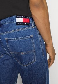 Mörkblå denimjeans med rak klippning, med en svart etikett med vit text och röda samt vita detaljer på baksidan.