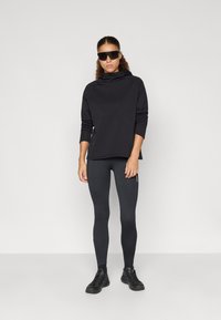 Μαύρο τοπ με μακριά μανίκια και ψηλό γιακά, συνδυασμένο με εφαρμοστά σκούρα κολάν και μαύρα αθλητικά παπούτσια. Το μοντέλο φοράει σκούρες ηλιό sunglasses.