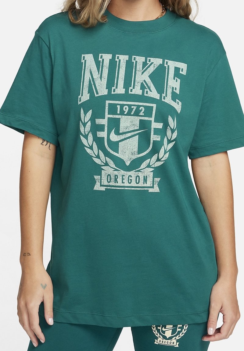 Turkusowa, oversize koszulka z bawełny z wyblakłym białym nadrukiem "NIKE 1972" oraz wzorem laurowym. Okrągły dekolt i krótkie rękawy.