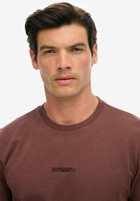 Mężczyzna z krótkimi ciemnymi włosami, ubrany w brązową koszulę Superdry, zwrócony przodem z neutralnym wyrazem twarzy na jednolitym jasnym tle.