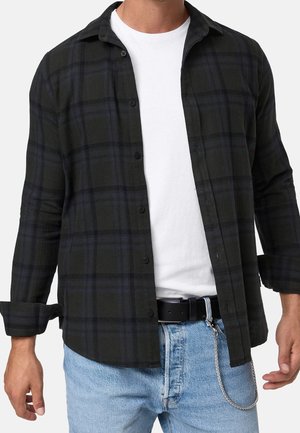 Mężczyzna ubrany w czarną i ciemnoniebieską koszulę w kratę zapinaną na guziki, na białej koszulce, jasnoniebieskie dżinsy i czarny pasek z srebrnym łańcuchem.