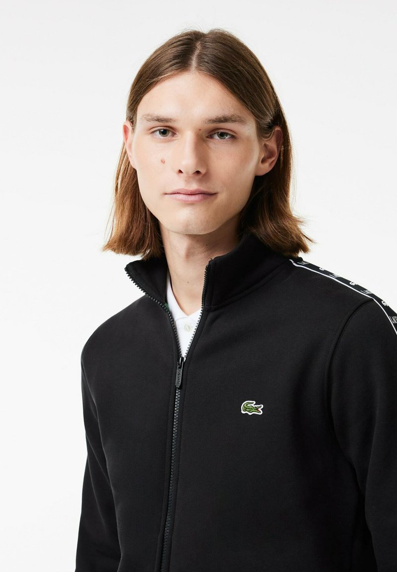 Schwarze Zip-Up-Jacke mit hohem Kragen, ausgestattet mit einem grünen gestickten Logo und weißen Streifen auf den Schultern. Weiche Stofftextur.