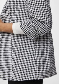 Camisa de patrón de espiga en blanco y negro con puños blancos, que presenta un corte relajado y una tela texturizada. Detalles de costura visibles.