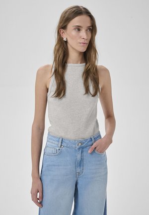 Młoda kobieta o długich brązowych włosach, ubrana w szary, bezrękawnik z prążkowanego materiału i jasnoniebieskie dżinsy z wysokim stanem, z ręką w kieszeni, na jednolitym tle.