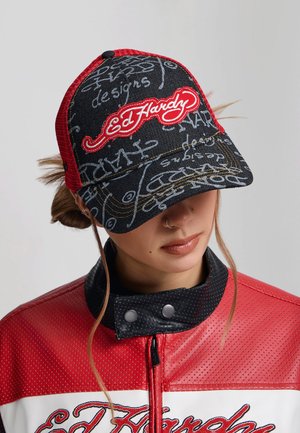 Młoda osoba nosząca czarno-czerwoną czapkę Ed Hardy oraz pasującą czerwoną skórzaną kurtkę, patrząca w dół z włosami schowanymi pod czapką.