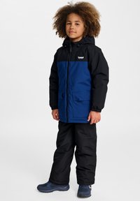 Une veste à capuche bleue et noire avec fermeture éclair à l'avant et deux grandes poches. L'enfant porte un pantalon noir et des chaussures bleues. Tissu texturé.