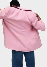 Blazer rose en tissu lisse, design double boutonnage avec un grand col et deux poches à l'avant. Porté sur un jean bleu.