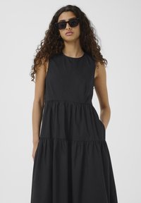 Schwarzes ärmelloses Kleid mit Rundhalsausschnitt und gestuften Design, das eine glatte Textur und seitliche Taschen aufweist. Model trägt Sonnenbrille.