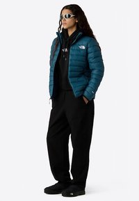 Türkisfarbene Steppjacke mit horizontalen Nähten, schwarzer Kapuzenpullover, lockere schwarze Hose und schwarze Schuhe. Sonnenbrille mit elegantem Design rundet den Look ab.