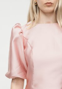 Ξανθιά γυναίκα που φορά ένα απαλό ροζ σατέν μπλουζάκι με φουσκωτά κοντά μανίκια και χρυσά σκουλαρίκια σε σχήμα σταγόνας, εμφανιζόμενη από τον λαιμό μέχρι τον άνω βραχίονα.