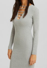 Langärmliges, geripptes graues Kleid mit tiefem V-Ausschnitt und mehreren horizontalen Ausschnitten. Figurbetonte Silhouette, weiche Textur.