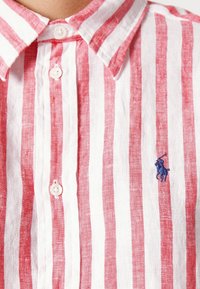 Camisa de botones a rayas rojas y blancas hecha de lino, con un logo bordado azul en el lado izquierdo del pecho y un diseño de cuello clásico.