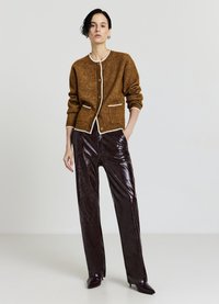 Braune Strickjacke mit weißen Borten und Knöpfen, kombiniert mit glänzenden burgunderroten Lederhosen. Beide Teile haben eine strukturierte Passform und Taschen.