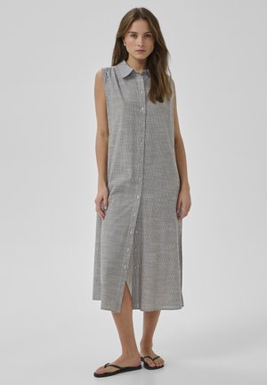 Nő ujjatlan, szürke-fehér csíkos midi ruhában, elől gombsorral és fekete papucsban, sima háttér előtt állva.