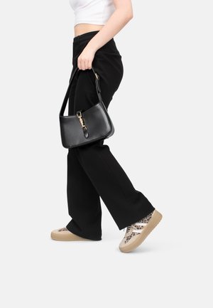 Zwarte leren handtas met een gouden sluiting, rechthoekige vorm, gedragen door iemand in zwarte broek en beigeruite sneakers.