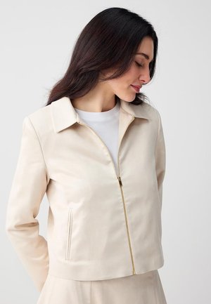 Femme aux longs cheveux foncés portant une veste crème à fermeture éclair sur un haut blanc, regardant vers le bas sur un fond clair uni.