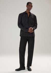 Camicia nera con bottoni e motivo testurizzato sottile, abbinata a pantaloni scuri e scarpe nere, per un look monocromatico. Sfondo semplice.