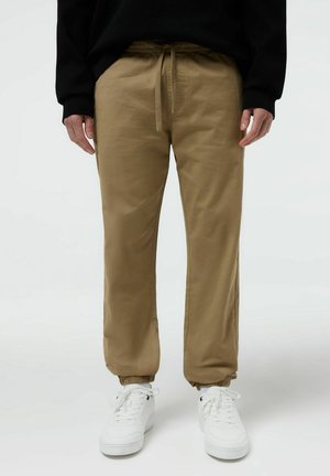 Mężczyzna w beżowych spodniach dresowych z elastycznymi ściągaczami i białych sneakersach, stojący na jednolitym białym tle.