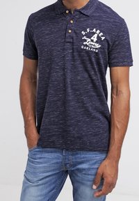 Navyblå polo skjorta med textilstruktur, knäppning med två knappar och vit tryckt design på vänster bröst. Matchad med ljusblå jeans.