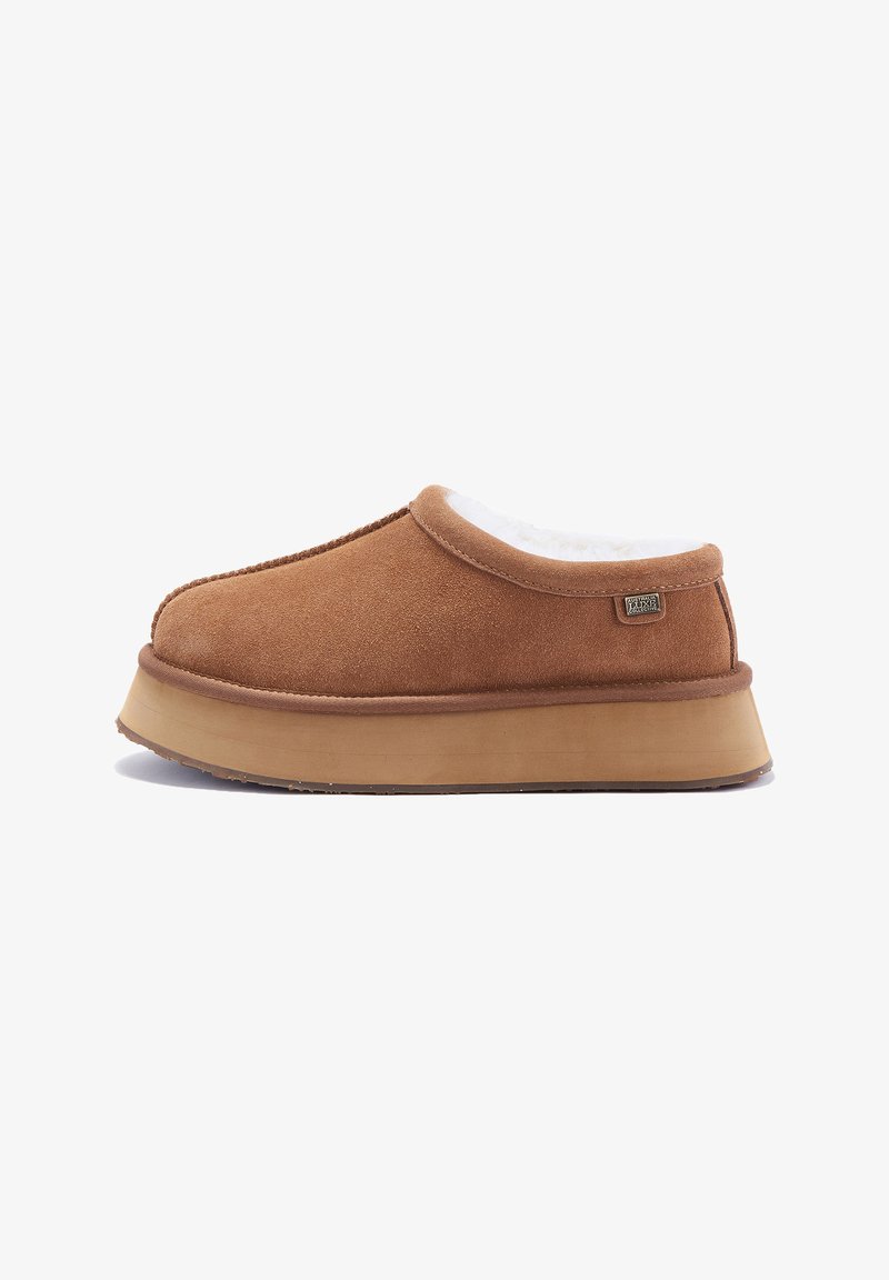 Chaussure à enfiler en daim marron avec un design arrondi, doublure en peluche blanche et semelle plate de couleur marron clair. Présente des coutures discrètes et une étiquette de logo.
