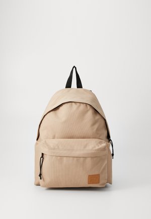 Mochila beige con un acabado textil suave, una parte superior redondeada, correas ajustables negras y un bolsillo delantero con cremallera que presenta detalles del logo.
