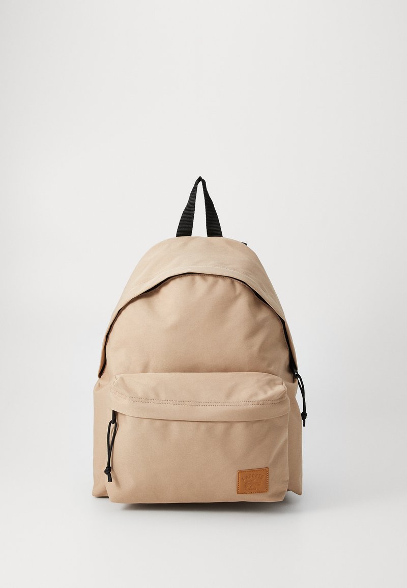Beige backpack with a smooth textile finish, a rounded top, adjustable black straps, and a front zipper pocket with logo detailing.