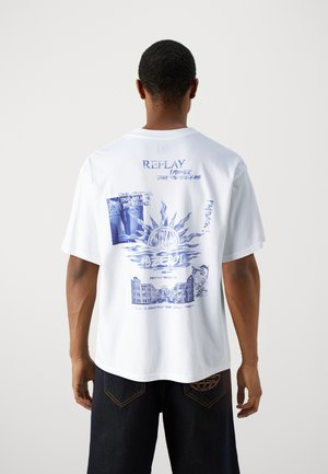 Mand iført hvid oversize t-shirt med blå grafiske designs og tekst på ryggen, kombineret med mørke denimshorts.