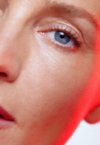 Gros plan d'un visage avec une peau lisse et claire, présentant un œil bleu saillant et une subtile lèvre rose. Une douce lumière rouge met en valeur les contours.