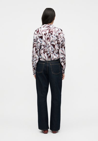 Γυναίκα που στέκεται με την πλάτη γυρισμένη, φορόντας ένα μακρυμάνικο πουκάμισο με λουλούδια, μπαίνει μέσα σε σκούρο μπλε τζιν και καφέ παπούτσια, σε απλό φόντο.