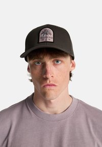 Jonge man met lichte sproeten, blauwe ogen en bruin haar, draagt een zwarte Billabong pet en een lichtpaars shirt met ronde hals.