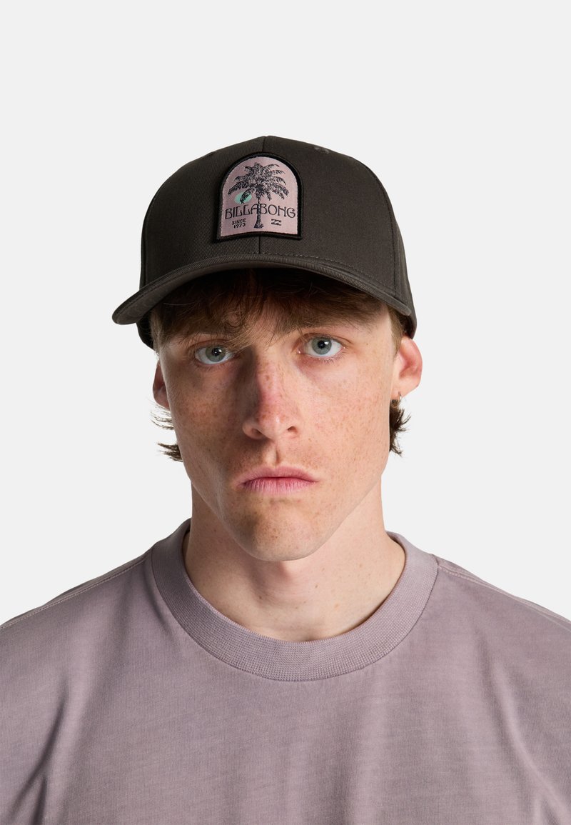 Jonge man met lichte sproeten, blauwe ogen en bruin haar, draagt een zwarte Billabong pet en een lichtpaars shirt met ronde hals.