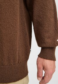 Brown textured sweater with ribbed cuffs and hem. Features a side slit and a small tag visible near the hem. Soft, knitted fabric.