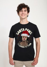 Svart t-shirt med ett motiv av en plyschhund som bär en röd tomteluva, med texten "SANTA PAWS ÄR PÅ VÄG TILL STAN" i vitt.