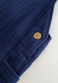 Tejido de punto azul marino con rayas verticales en azul más claro. Presenta un botón de madera redondo y una superficie texturizada.