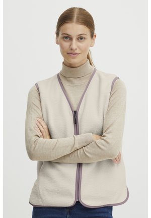 Gilet en polaire beige avec une fermeture éclair à l'avant, bordé de violet clair, présentant un ourlet arrondi et deux poches latérales. Porté par-dessus un col roulé beige.
