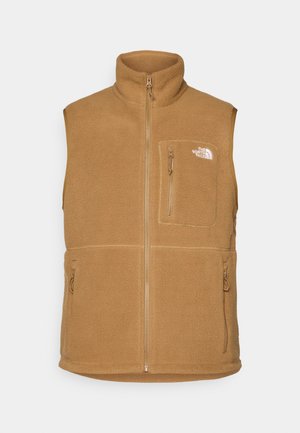 Tan fleece vest met hoge kraag, ritssluiting aan de voorkant en twee zijzakken. Bevat een borstzak en een logo aan de linkerkant. Zachte textuur.