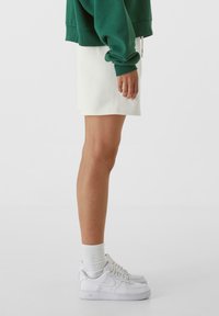 Bílá mini sukně a zelená oversized mikina, doplněné bílými teniskami a žebrovanými ponožkami. Jednoduchý design, neformální vzhled, minimalistický styl.