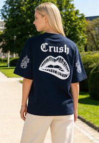 Donna bionda in piedi all'aperto che indossa una maglietta nera con la scritta bianca "Crush" e una grande grafica di labbra sulla schiena.