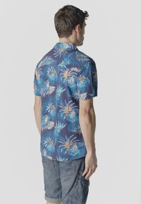 Hombre con camisa hawaiana azul de manga corta con patrones florales y pantalones cortos cargo de mezclilla, visto de espaldas contra un fondo liso.