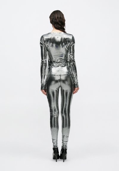 Hõbedase litritega kombinesoon, millel on pikad varrukad, keha järgnev lõige ja kõrge krae. Helkiva tekstuuriga ja kitsenevate sääreosadega. Kandmiseks koos kõrgete kontsadega.