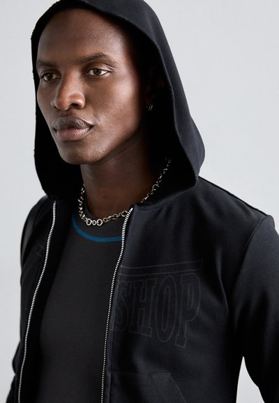 Black zip-up hooded jacket with front pockets, featuring a subtle "SHOP" print. Textured fabric with a smooth finish; layered over a ribbed gray tank.
