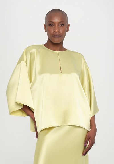 Blusa de satén de manga corta en amarillo claro con escote redondo y detalle de ojo de cerradura. Corte relajado y textura suave.