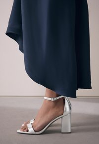 Sandales à talons hauts argentées avec un talon bloc, une bride de cheville et deux brides à l'avant. Posées sur une jupe fluide bleu marine.