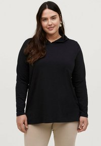 Sort langærmet hættebluse med en rund halsudskæring og afslappet pasform, lavet af blødt stof, båret med beige bukser. Minimalistisk design.