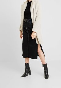 Manteau en fausse fourrure crème, col roulé noir, jupe en denim noir taille haute avec fente latérale, et bottines à talons noires.